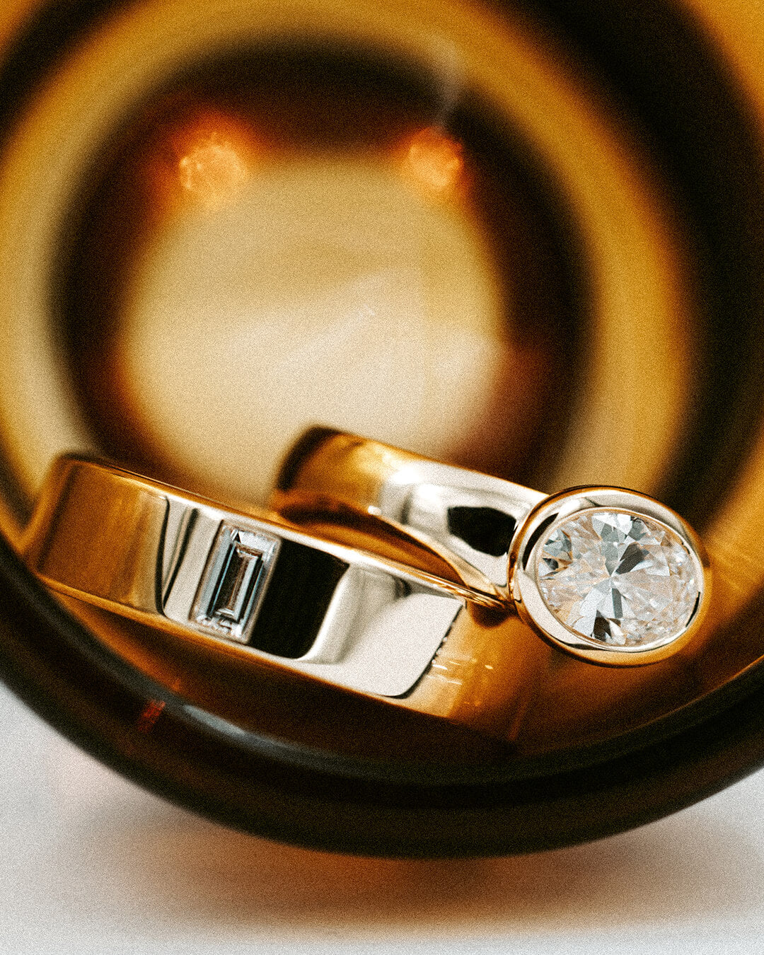 A close-up shot of two wedding bands, one yellow gold with a round brilliant-cut diamond and the other with a rectangular baguette-cut diamond, resting inside a dark amber glass bowl. Custom jewellery designed in New Zealand by BY TONIN.