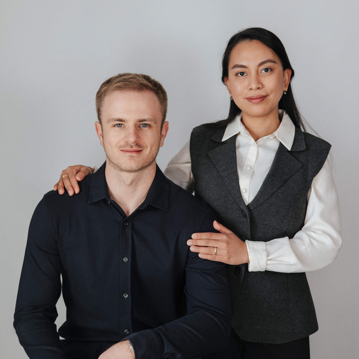 Founders of BY TONIN Jewellery based in NZ. Carlo Tonin in a dark navy blue shirt and Mia in a dark gray vest over a white collared shirt, posing together against a light gray studio background.