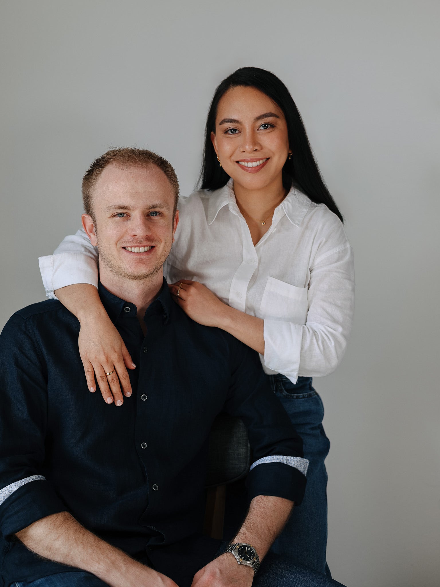 A professional portrait of Carlo and Mia Tonin, the founders of BY TONIN. Carlo, a smiling man with short blonde hair, sits in a chair wearing a dark blue button-down shirt with patterned cuffs. Mia, a smiling woman with long dark hair, stands behind him with her arms resting on his shoulders. She is wearing a white linen button-down shirt and denim jeans. The background is a simple, light gray wall.