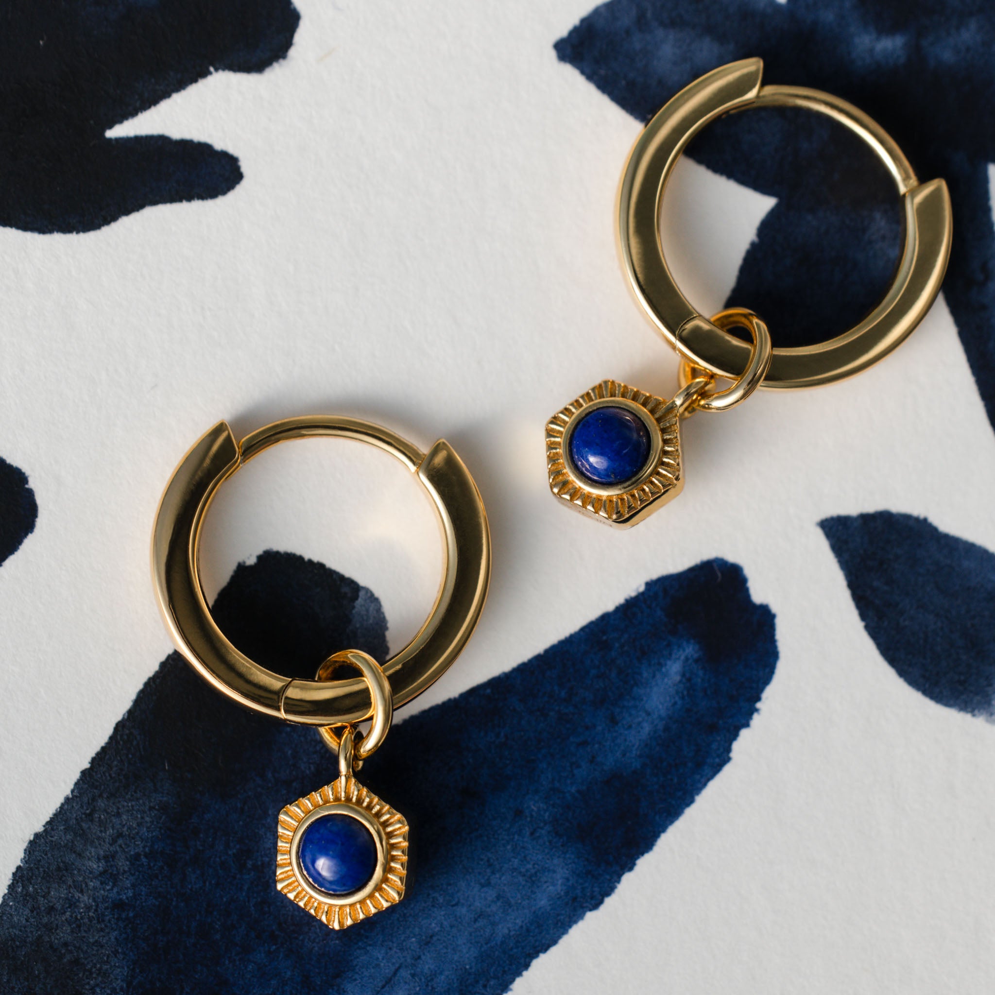 Make a captivating statement with BY TONIN Bel e Viv gold hoop earrings, featuring vibrant hexagonal lapis lazuli charms, artfully presented on a dynamic blue patterned background.