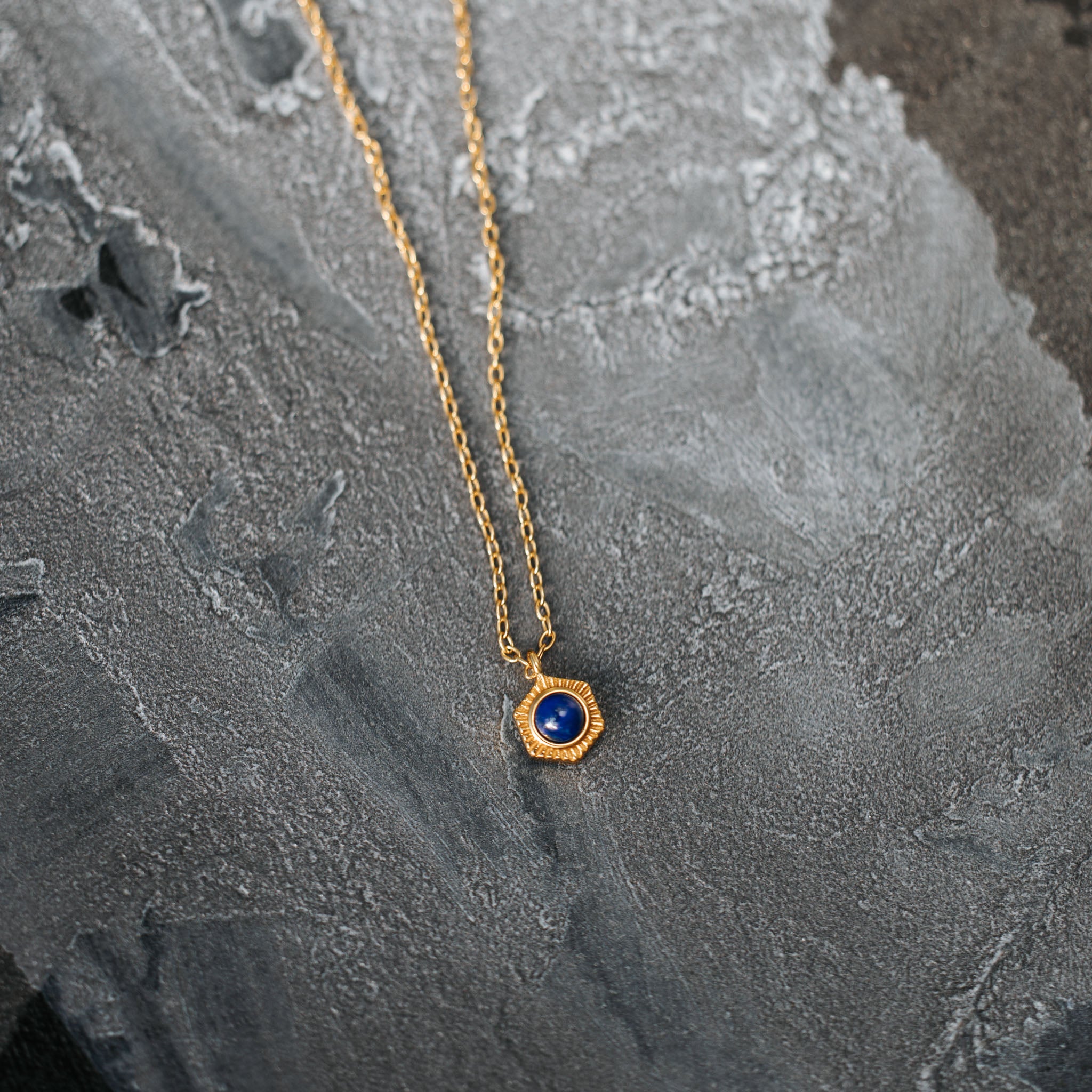 Command attention with BY TONIN's Bel e Viv gold necklace, featuring a mesmerising hexagonal lapis lazuli pendant, dramatically presented on a textured grey surface.