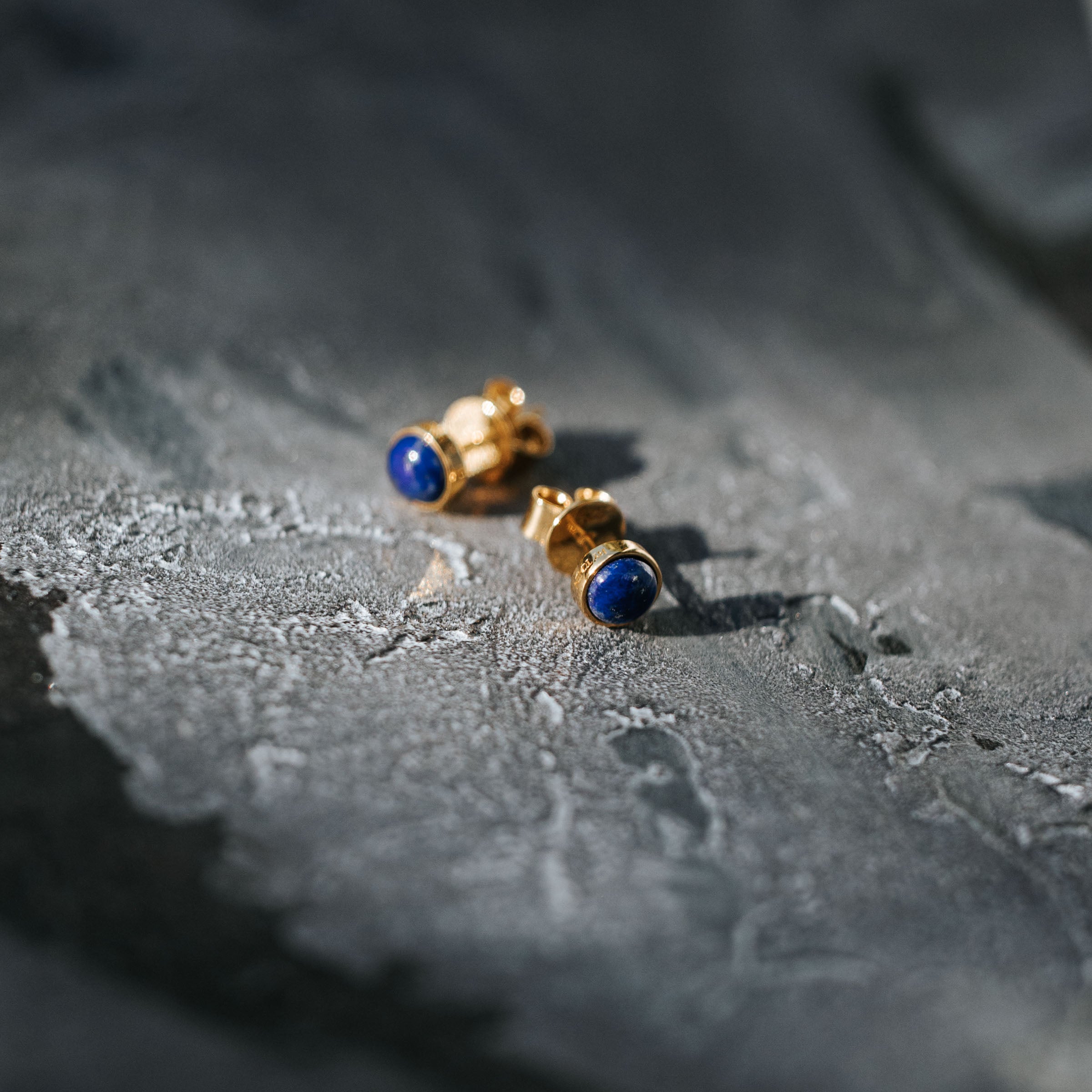 Illuminate your style with BY TONIN's Bel e Viv gold round stud bezel earrings, featuring captivating deep blue lapis lazuli, artfully presented on a textured grey surface.