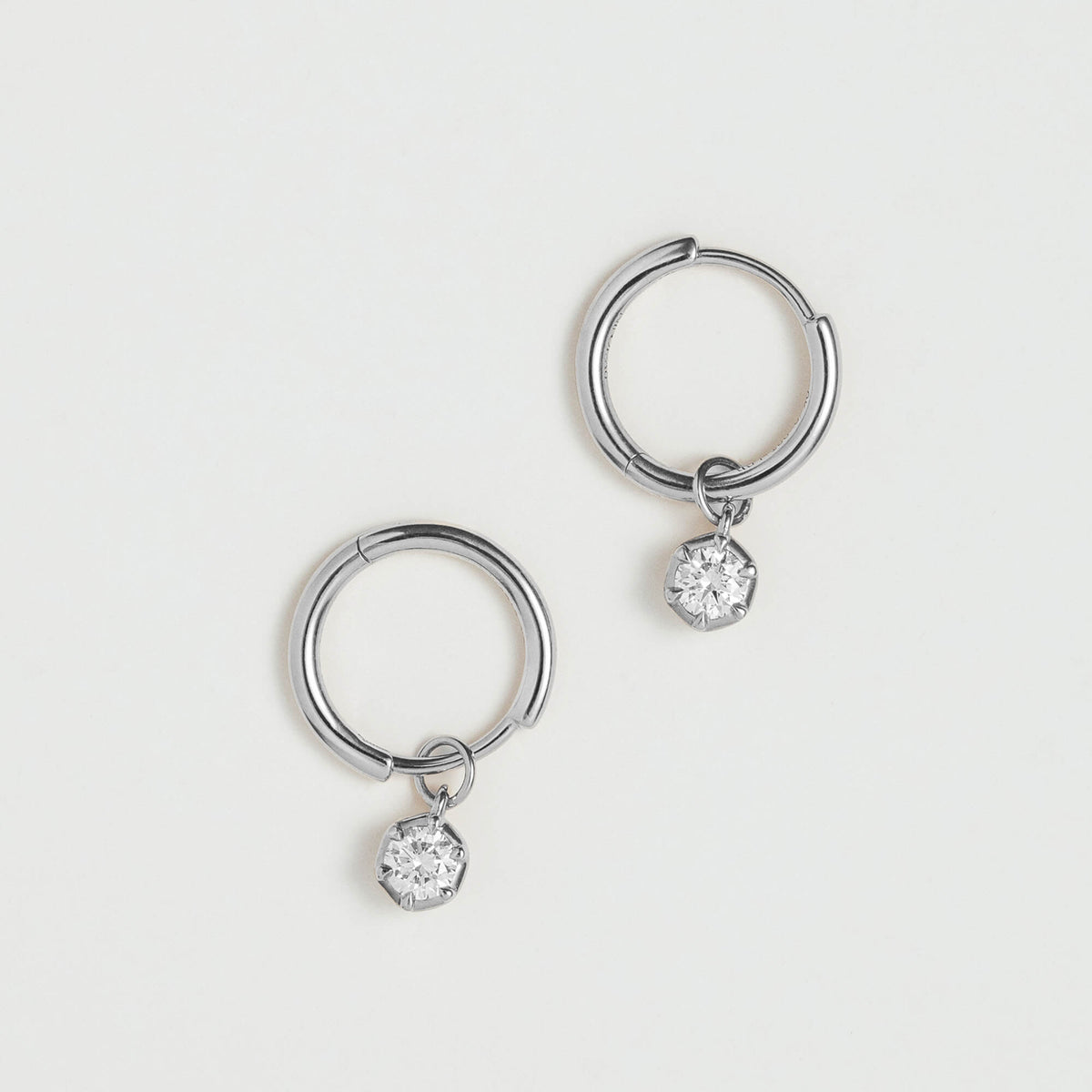 A pair of silver-toned hoop earrings, each with a small clear faceted gemstone dangling from the bottom, displayed on a clean off-white background.