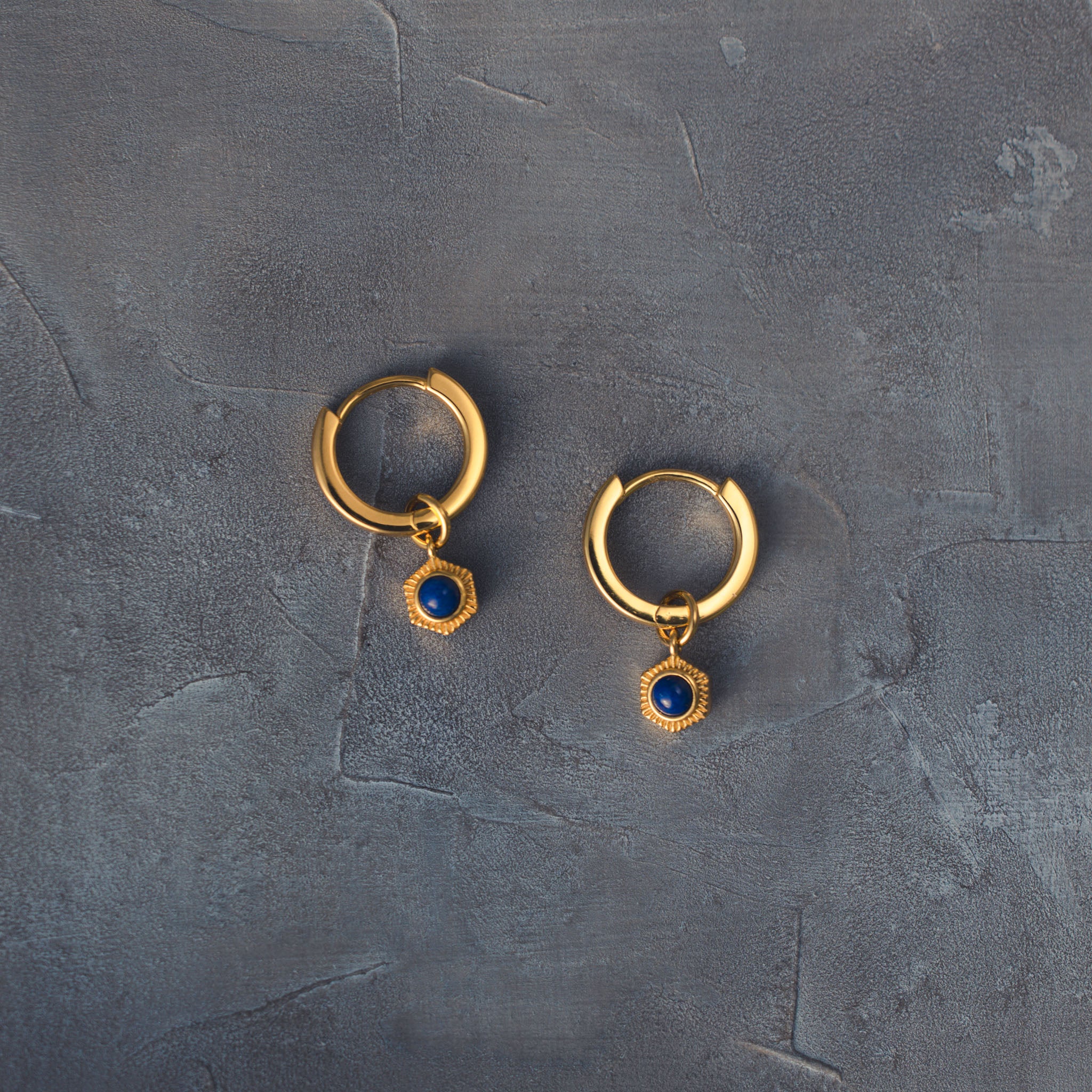Adorn your ears with BY TONIN's radiant gold hoop earrings, featuring captivating deep blue lapis lazuli drop charms, elegantly presented on a dark grey textured surface.