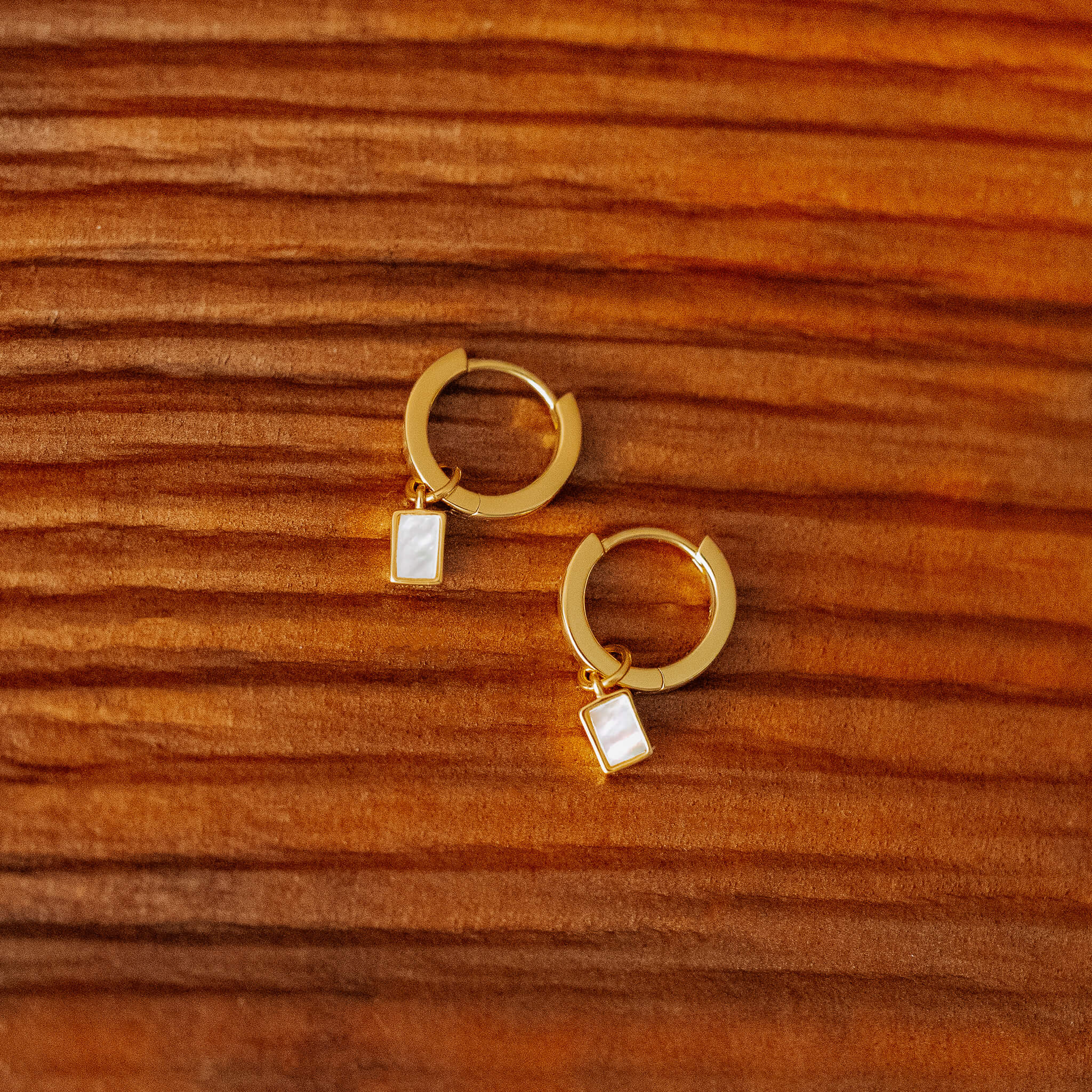 A pair of gold hoop earrings with small, rectangular mother-of-pearl charms dangling from them, resting on a wooden surface.