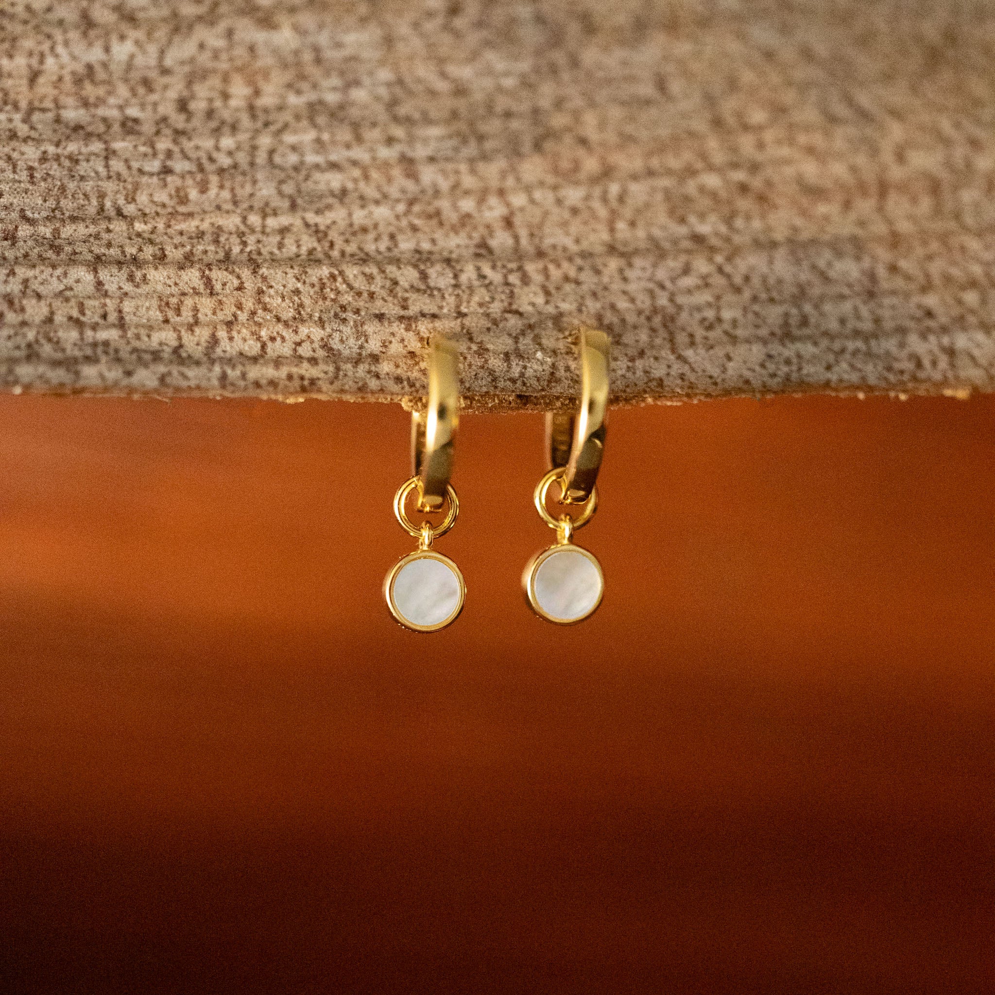 Embrace timeless elegance with BY TONIN's Heritage gold hoop earrings, featuring luminous round mother of pearl charms, gracefully displayed on a rich wooden surface.