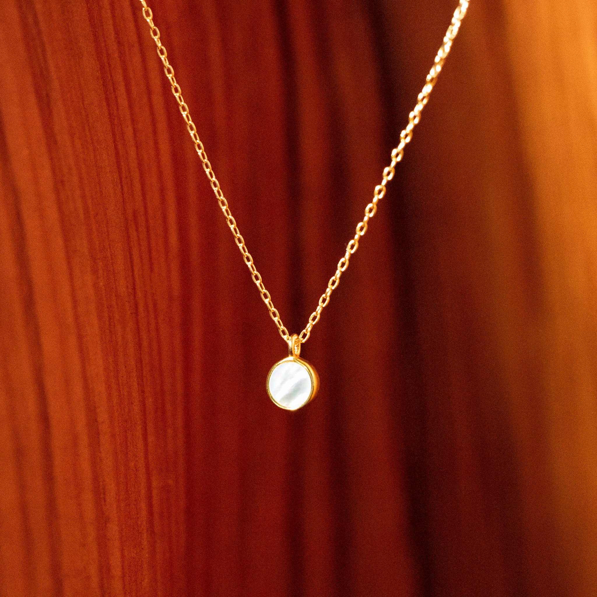 Embrace the timeless glow of BY TONIN's Heritage gold necklace, showcasing a luminous round mother of pearl pendant, gracefully displayed on a rich wooden surface.