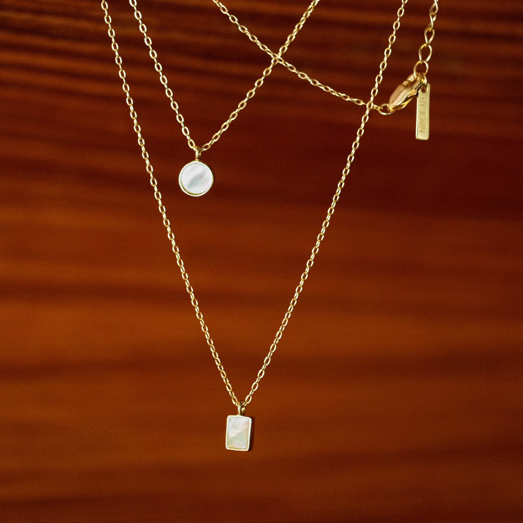 Master the art of elegant layering with BY TONIN's exquisite gold necklaces, featuring a luminous round mother of pearl pendant paired with a chic rectangular design, beautifully displayed on a rich brown surface.