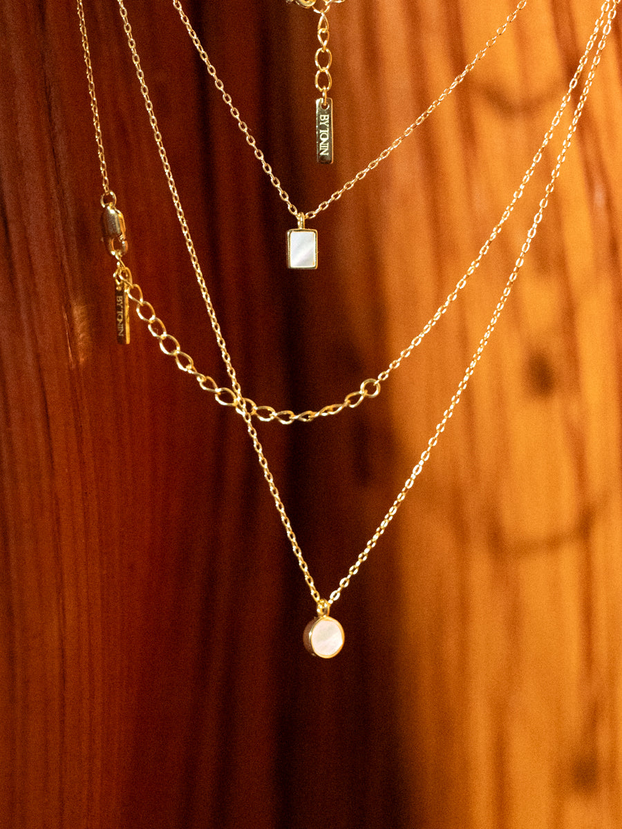 Master the art of sophisticated layering with BY TONIN's exquisite gold necklaces, featuring luminous round and rectangular mother of pearl pendants, elegantly draped on a rich wooden surface.