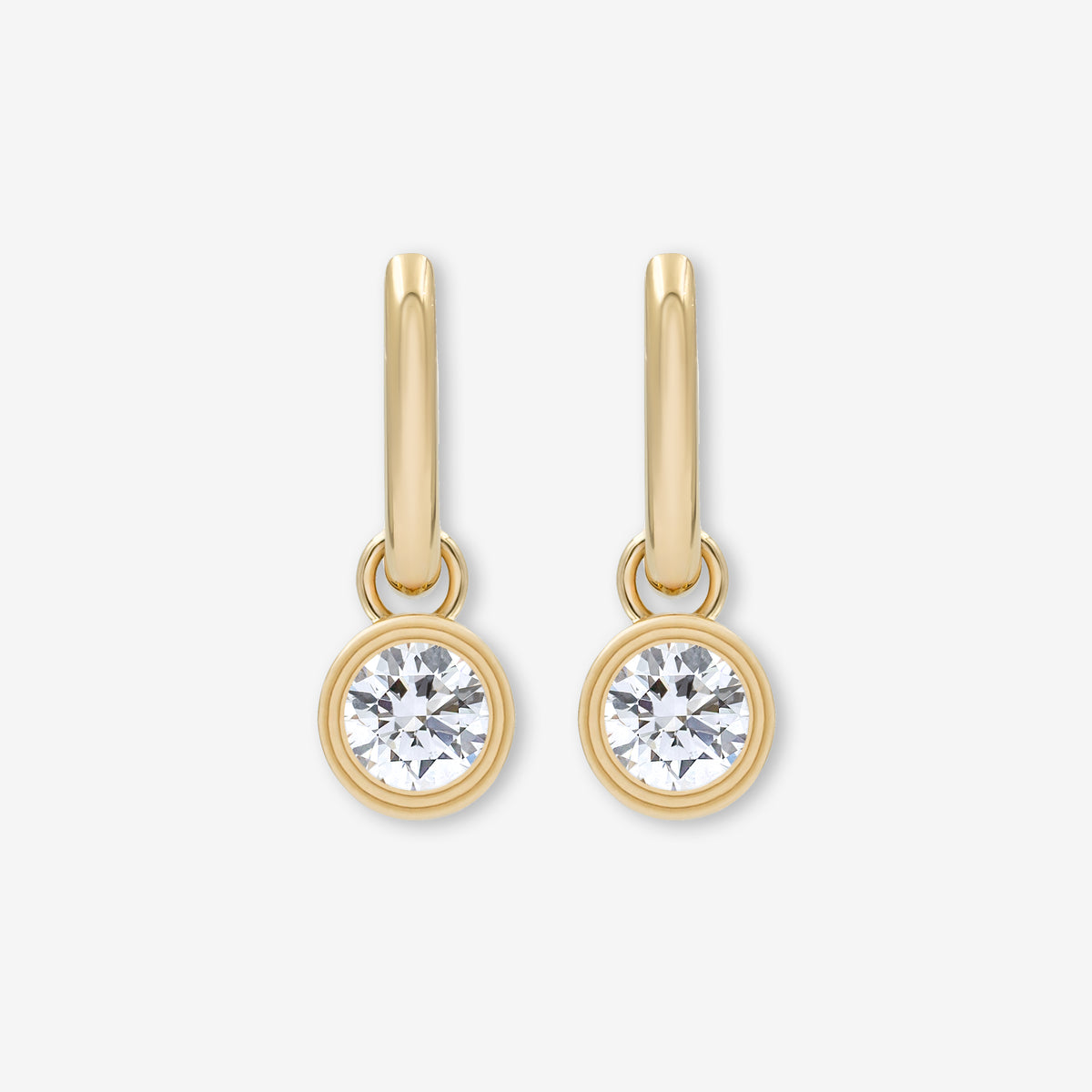 A pair of gold drop earrings with a single round clear lab grown diamond in a bezel setting, shown against a light gray background.