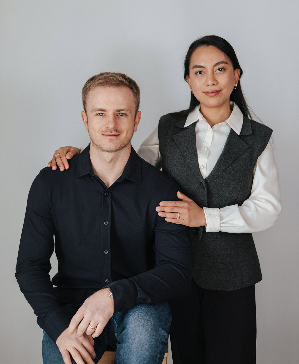 Founders of BY TONIN Jewellery based in NZ. Carlo Tonin in a dark navy blue shirt and Mia in a dark gray vest over a white collared shirt, posing together against a light gray studio background.