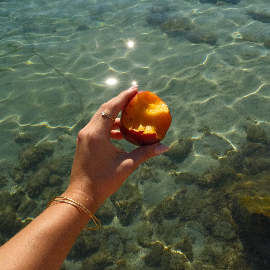 A hand holding a partially eaten peach fruit, wearing a ring with baguette mother of pearl over clear, sunlit water with rocks visible beneath the surface.