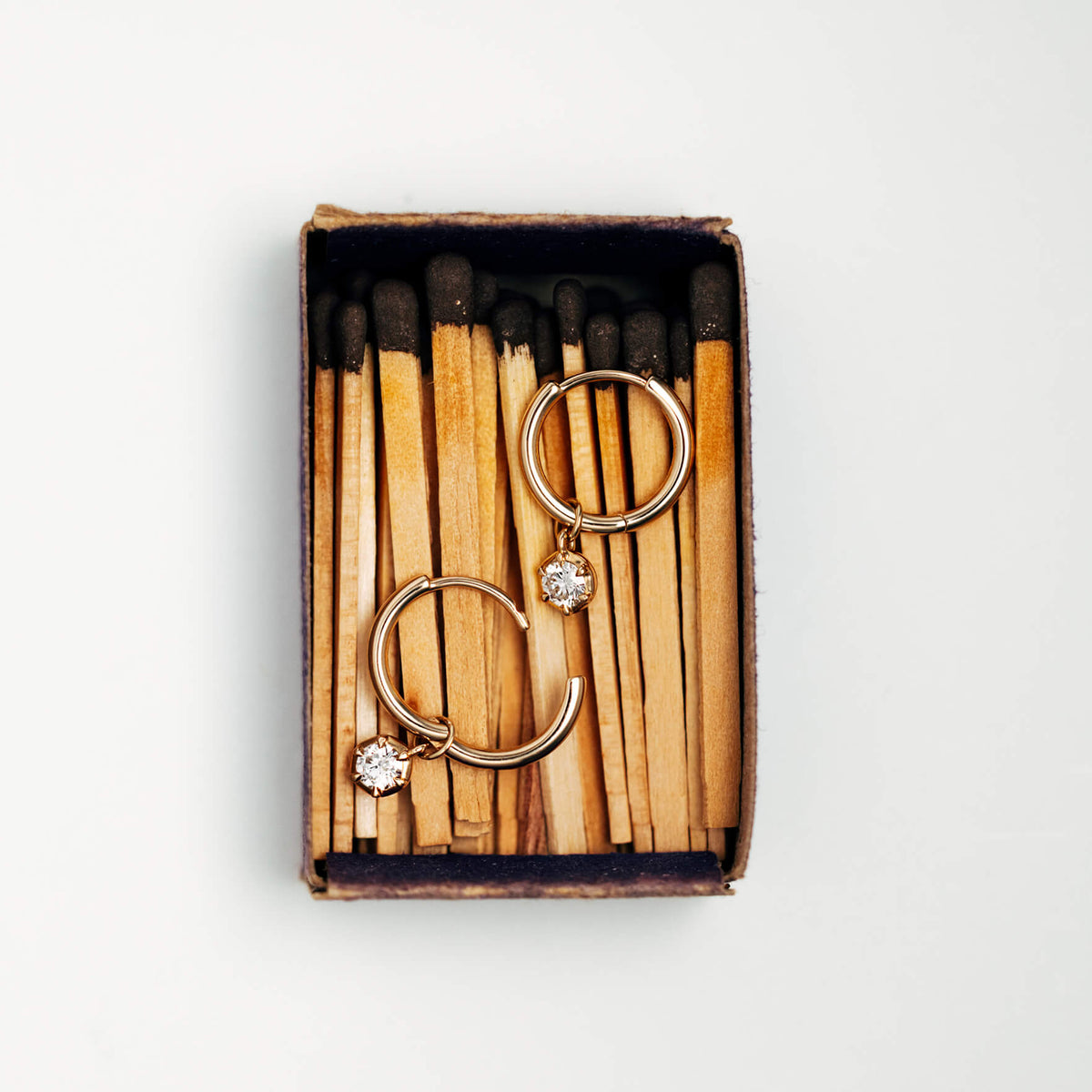 Gold hoop earrings with clear diamond accents, displayed inside an open vintage-style matchbox filled with wooden matches on a light gray background.