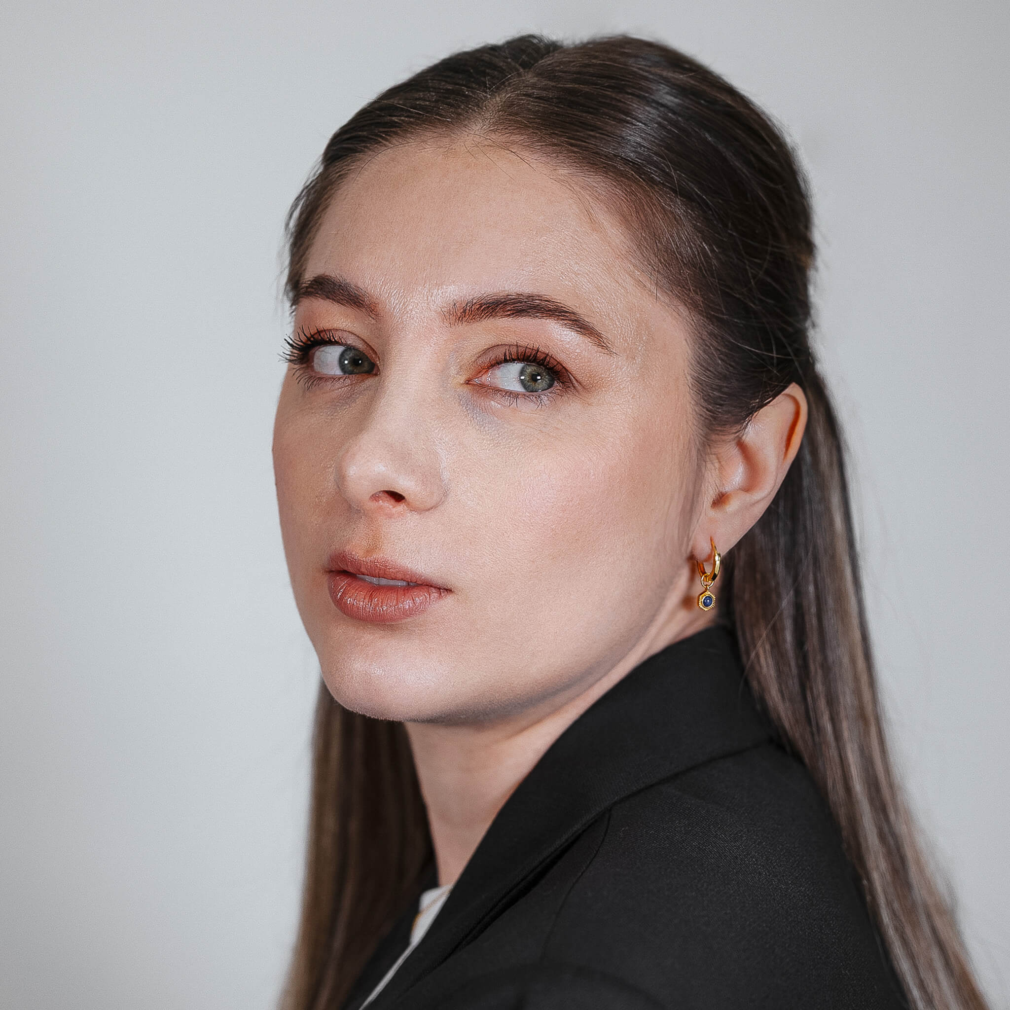 Woman with long hair wearing a gold hoop earrings with lapis lazuli, black blazer against a plain background