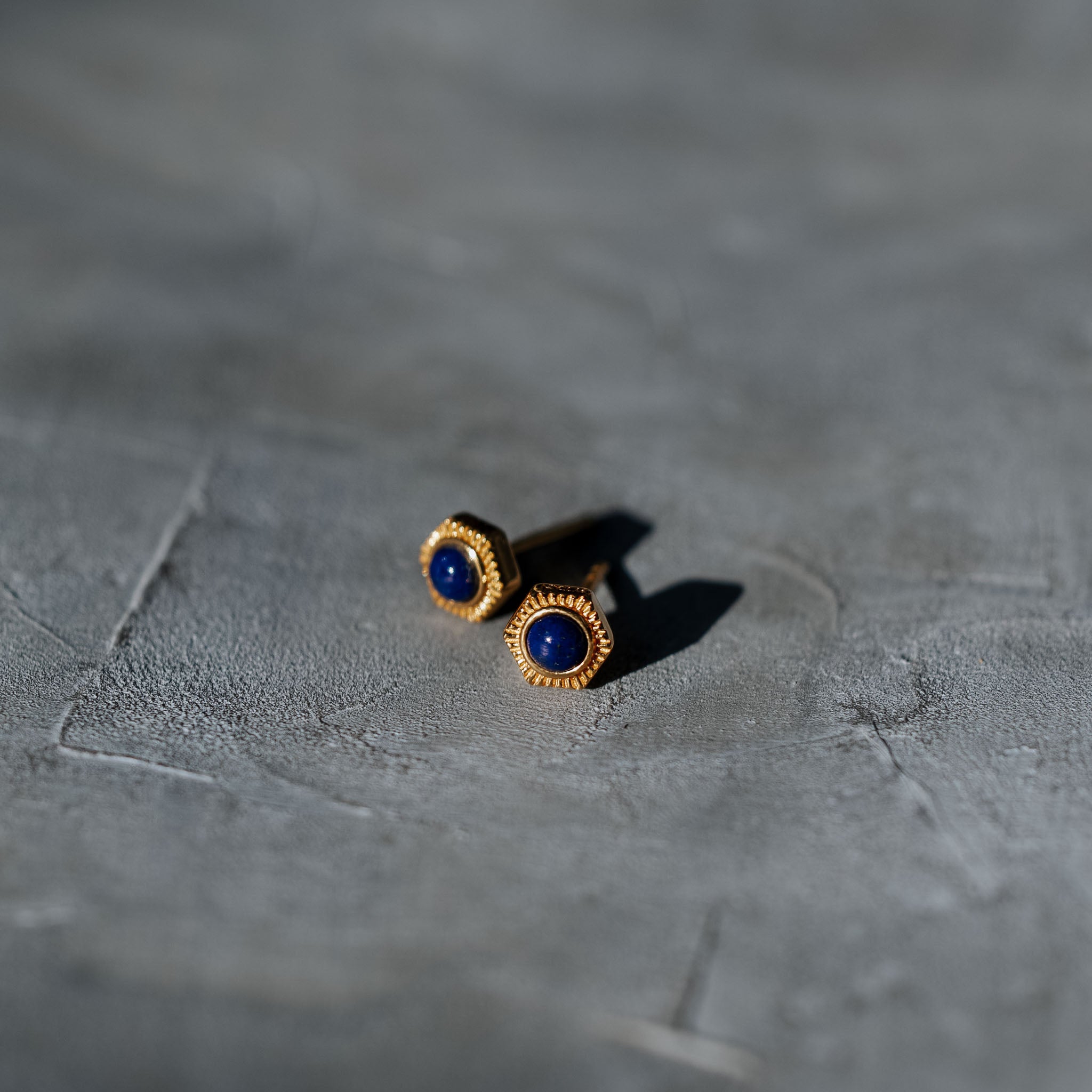 Illuminate your style with BY TONIN's Bel e Viv gold round hexagon earrings, featuring captivating deep blue lapis lazuli, artfully presented on a textured grey surface.