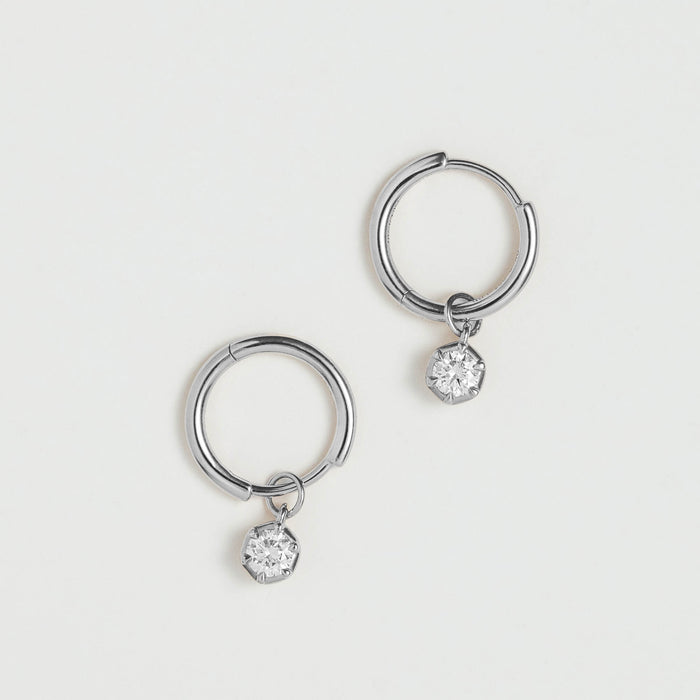 A pair of silver-toned hoop earrings, each with a small clear faceted gemstone dangling from the bottom, displayed on a clean off-white background.