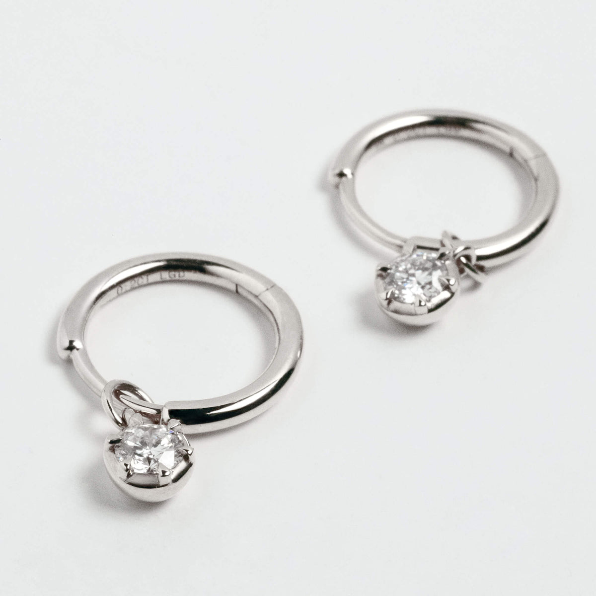 A pair of silver-toned huggie hoop earrings, each featuring a small clear diamonds in a prong setting, displayed on a clean white background.