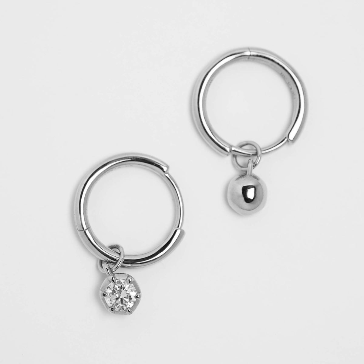 A pair of silver hoop earrings, each with a small dangling charm. One charm is a smooth polished back, and the other is a round clear diamond in a prong setting.