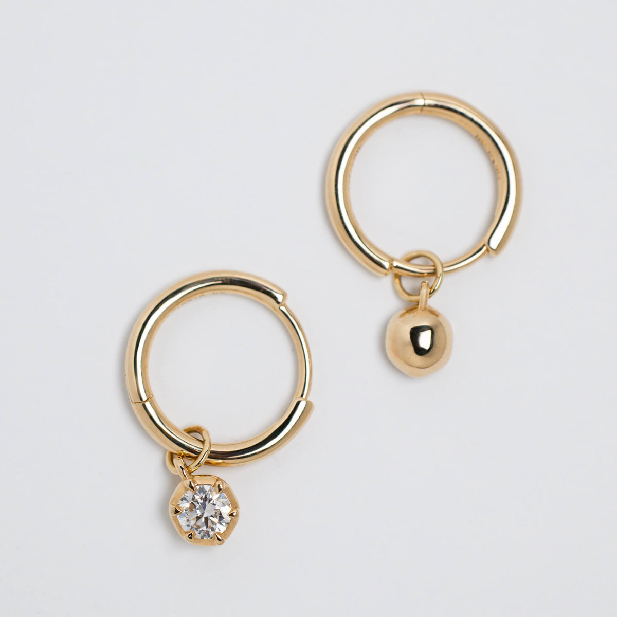 A pair of gold hoop earrings, one with a solid gold back and the other with a lab grown diamond, shown on a light gray background.