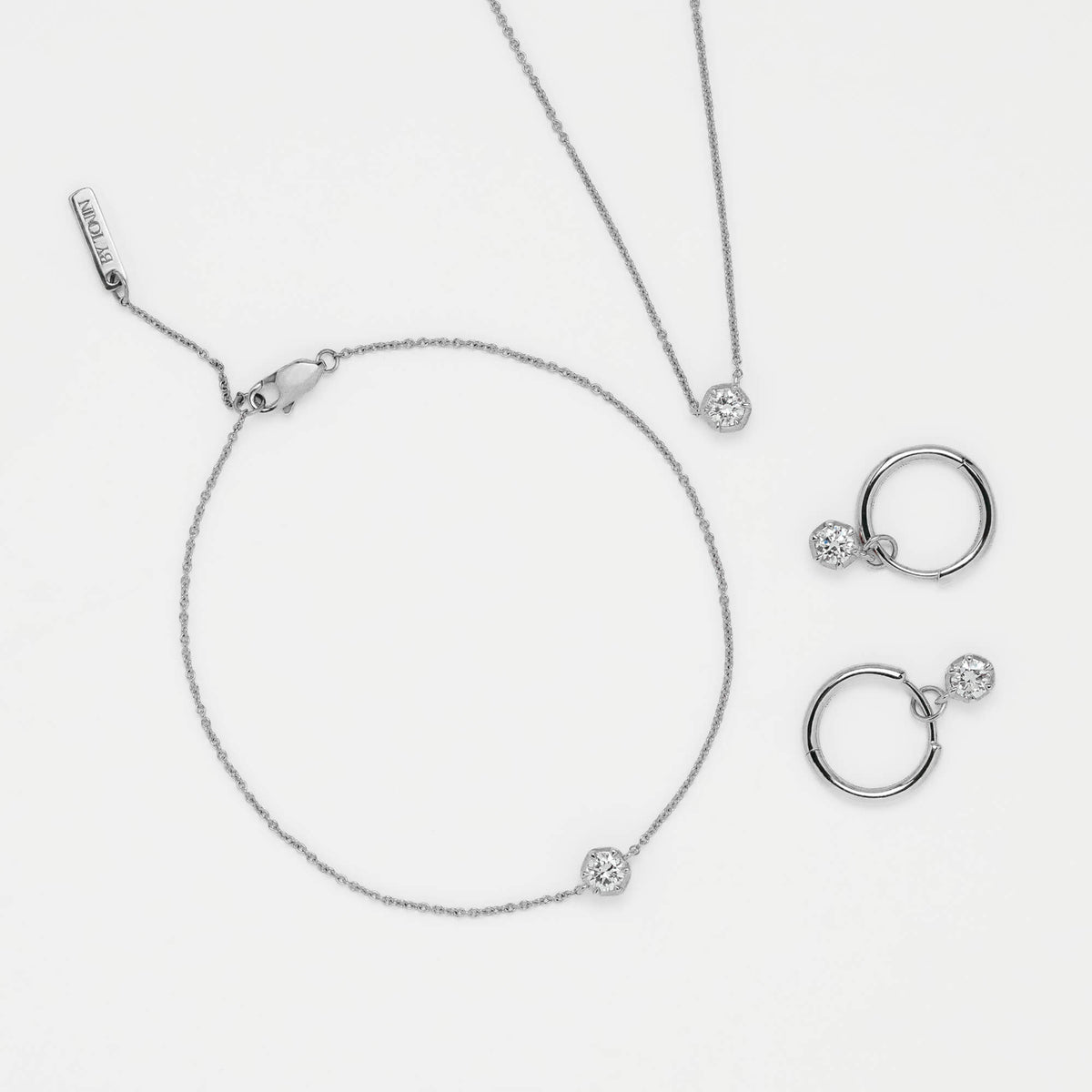 A silver jewelry set featuring a delicate chain bracelet with a small round bezel-set stone, a matching necklace, and a pair of hoop earrings with small dangling beads and round stones, all arranged on a clean white background.