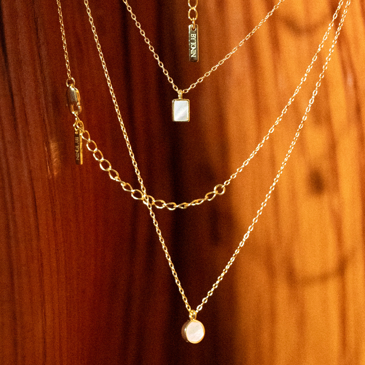 Master the art of sophisticated layering with BY TONIN's exquisite gold necklaces, featuring luminous round and rectangular mother of pearl pendants, elegantly draped on a rich wooden surface.
