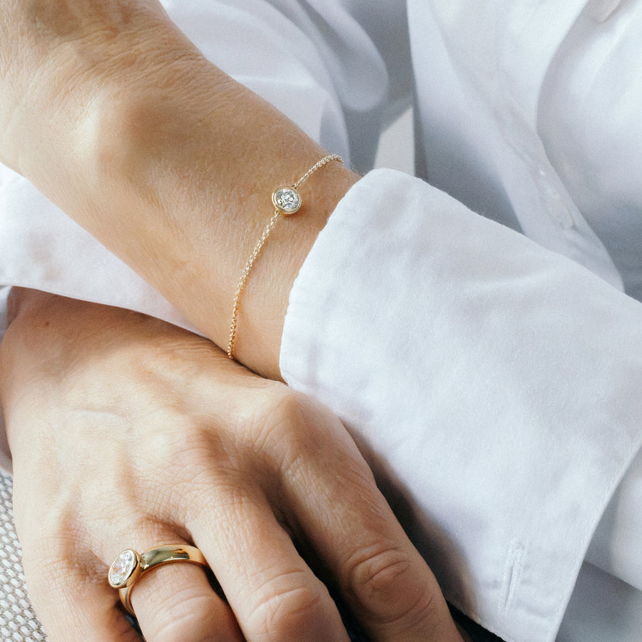 A delicate gold chain bracelet with a small round clear diamond pendant, worn on a person's wrist. The person is wearing a white button-down shirt, with their hands resting in the foreground.